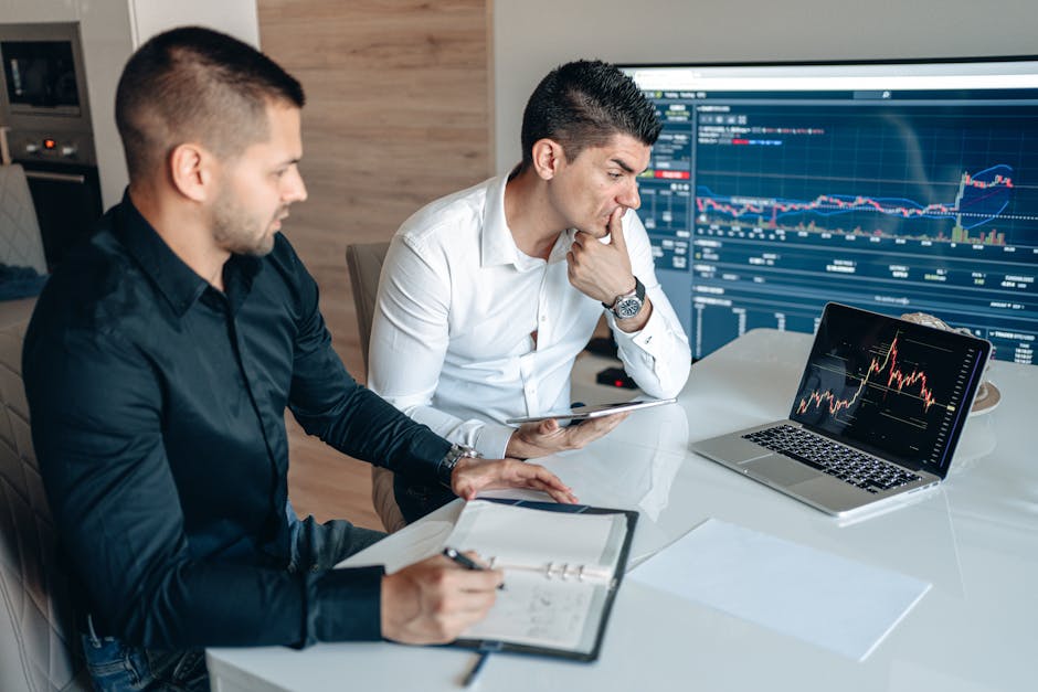 Two businessmen sitting at a desk analyzing stock charts on laptops and tablets.
